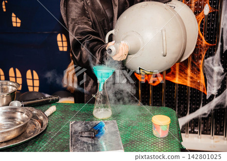 Person in wizard black robe pouring liquid nitrogen from a large dewar tank into a glass flask using a funnel during a scientific show or Halloween theme party. Steaming reaction experiment with smoke 142801025
