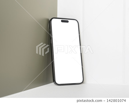 Phone mockup with a blank white screen leans against a grey interior wall corner. Template display Phone mockup with a blank white screen leans against a grey interior wall corner. Template display 142801074