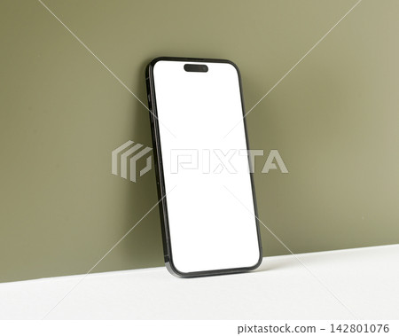 Phone mockup with a blank white screen leans against an olive-toned interior wall, photographed at Phone mockup with a blank white screen leans against an olive-toned interior wall, photographed at 142801076