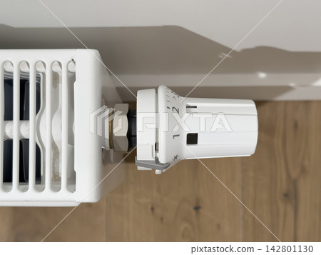 Thermostatic radiator valve on a white heater. Close up heating control in a home interior. Energy saving temperature regulation for winter comfort.. Thermostatic radiator valve on a white heater. Close up heating control in a home interior. Energy saving temperature regulation for winter comfort.. 142801130