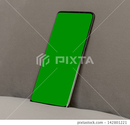 Smartphone mockup with green screen on a gray fabric background. Angled mobile phone leans against a cushion in a clean interior setup. Ideal for app design, advertising, and 142801221