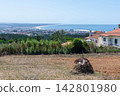 Scenic panoramic landscape view of a coastal town in Portugal from a rustic hill. A haystack sits in the foreground field under a clear blue sky. Tranquil summer vibe, nobody. 142801980