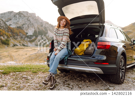 Young woman enjoying autumn nature while sitting in the trunk of her car during a trip. Road trip. Young woman enjoying autumn nature while sitting in the trunk of her car during a trip. Road trip. 142802116