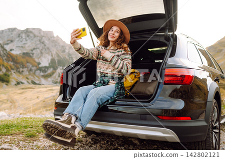 Selfie by car traveling. Young woman using a smartphone while sitting in the trunk of car in nature. Selfie by car traveling. Young woman using a smartphone while sitting in the trunk of car in nature. 142802121