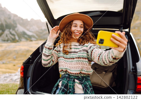Selfie by car traveling. Young woman using a smartphone while sitting in the trunk of car in nature. Selfie by car traveling. Young woman using a smartphone while sitting in the trunk of car in nature. 142802122