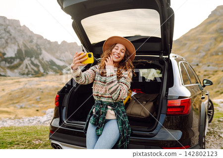 Selfie by car traveling. Young woman using a smartphone while sitting in the trunk of car in nature. Selfie by car traveling. Young woman using a smartphone while sitting in the trunk of car in nature. 142802123