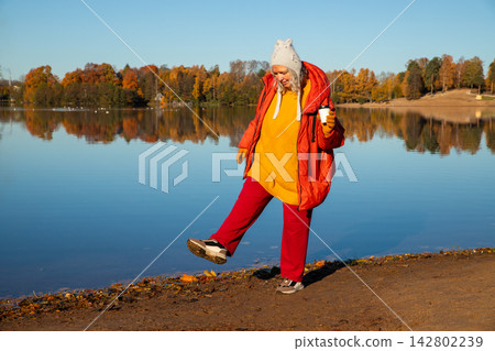 Mature woman walking along a pond shore in autumn. Happy middle aged lady enjoys outdoor leisure time with hot drink. Lifestyle clothing concept for seasonal nature walk activity. 142802239