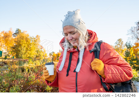 Woman hiker in autumn forest holding disposable paper coffee cup. Mature forty seven year old lady relaxing during trekking trip. Bodypositive active lifestyle in outdoor environment. Woman hiker in autumn forest holding disposable paper coffee cup. Mature forty seven year old lady relaxing during trekking trip. Bodypositive active lifestyle in outdoor environment. 142802246