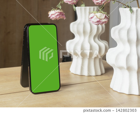 Smartphone mockup with a blank white screen leans on a wooden table near decorative ceramic vases Smartphone mockup with a blank white screen leans on a wooden table near decorative ceramic vases 142802303