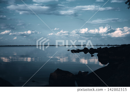Moody waterfront landscape with calm, reflective water, dark shoreline rocks, dramatic clouds and soft blue evening light, creating serene atmospheric nature scene for travel, wellness and design use. 142802315