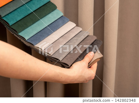 Fabric sample book held near window curtain in showroom. Designer selecting soft grey upholstery Fabric sample book held near window curtain in showroom. Designer selecting soft grey upholstery 142802348