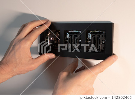 Power socket outlet frame plate installation on white wall. Hands placing black cover unit over Power socket outlet frame plate installation on white wall. Hands placing black cover unit over 142802405