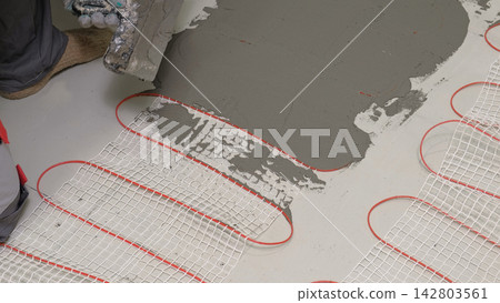 Installation of electric underfloor heating system with mortar tile adhesive close up. Home renovation process. Floor heating mat placement under cement screed layers for DIY house remodeling concept. Installation of electric underfloor heating system with mortar tile adhesive close up. Home renovation process. Floor heating mat placement under cement screed layers for DIY house remodeling concept. 142803561