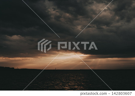 Dramatic storm clouds part above a dark horizon as warm sunbeams break through, creating an atmospheric landscape with golden light, powerful contrast and moody natural sky scene. Cloudscape view. 142803607