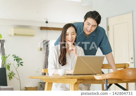 一位年輕的日本女性在家裡的桌子旁使用筆記型電腦工作,一位年輕的日本男性在一旁看著她。 一位年輕的日本女性在家裡的桌子旁使用筆記型電腦工作,一位年輕的日本男性在一旁看著她。 142804731