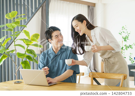 一對年輕的日本明星夫婦:一位男士在家裡的桌子旁用筆記型電腦工作,一位女士手捧一杯水。 一對年輕的日本明星夫婦:一位男士在家裡的桌子旁用筆記型電腦工作,一位女士手捧一杯水。 142804733