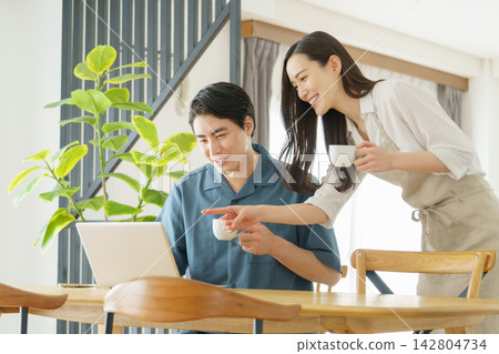 一對年輕的日本明星夫婦:一位男士在家裡的桌子旁用筆記型電腦工作,一位女士手捧一杯水。 一對年輕的日本明星夫婦:一位男士在家裡的桌子旁用筆記型電腦工作,一位女士手捧一杯水。 142804734