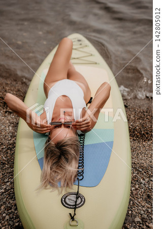 Blonde woman relaxing on paddleboard wearing white swimsuit during sunny day at lake shore 142805012