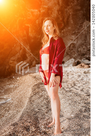 Woman beach vacation mature female in red bikini and shirt standing on pebble shore enjoying golden hour sunset by the sea 142805040