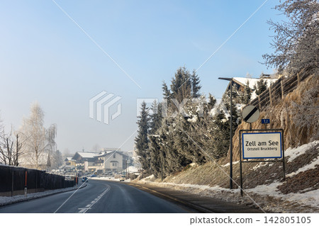 Winter road enters Zell am See Bruckberg in Austria, passing snowy slopes, evergreen trees and alpine homes under clear blue sky. Roadside sign marks resort destination amid frosty mountain scenery 142805105