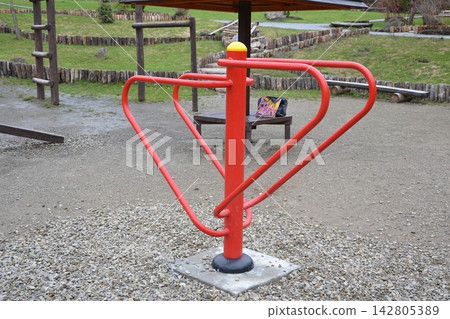 Red metal outdoor sports equipment for physical exercise and waist twister training, set on gravel ground in a public park street gym. Background features a resting area with wooden benches, ladder Red metal outdoor sports equipment for physical exercise and waist twister training, set on gravel ground in a public park street gym. Background features a resting area with wooden benches, ladder 142805389