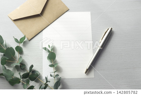 A wood-grain background with a kraft envelope, white stationery, a pen, and eucalyptus leaves arranged on it. A wood-grain background with a kraft envelope, white stationery, a pen, and eucalyptus leaves arranged on it. 142805782