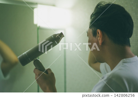 The back view of a man in his late 30s drying his hair with a hairdryer in a changing room at night. 142806256