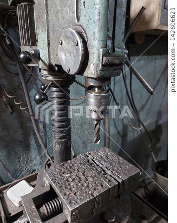 Close-up of a vintage industrial drill press with metal vise and drill bit in an old workshop. Worn machinery, rust, grease, and heavy-duty equipment create an authentic manufacturing and engineering Close-up of a vintage industrial drill press with metal vise and drill bit in an old workshop. Worn machinery, rust, grease, and heavy-duty equipment create an authentic manufacturing and engineering 142806621