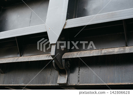 Close-up view of the heavy-duty black painted metal locking mechanism and hinge structure of a dump truck or tipper trailer body, highlighting industrial engineering, steel construction, and heavy Close-up view of the heavy-duty black painted metal locking mechanism and hinge structure of a dump truck or tipper trailer body, highlighting industrial engineering, steel construction, and heavy 142806628