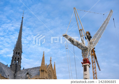 Final stage of Notre-Dame Cathedral of Paris reconstruction, part of building with church spire and roof and crane boom against blue sky. Renovation of historical landmark after devastating fire Final stage of Notre-Dame Cathedral of Paris reconstruction, part of building with church spire and roof and crane boom against blue sky. Renovation of historical landmark after devastating fire 142806885