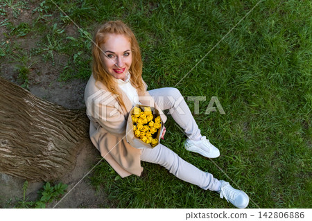 Red haired woman sitting on vibrant green grass next to a tree trunk, holding a beautiful bouquet of yellow tulips, smiling directly at the viewer and embracing the joyous essence of nature 142806886