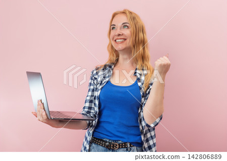 Joyful woman holding a notebook computer and making a winning gesture, experiencing online achievement or conceptualizing a new idea, isolated on a pink background 142806889