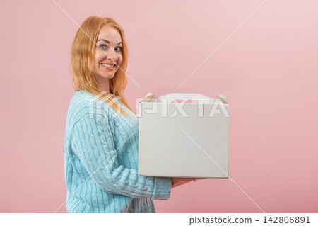 Smiling red haired woman holding a blank white cardboard carry box with a handle, presenting a delivery or a gift against a pink background Smiling red haired woman holding a blank white cardboard carry box with a handle, presenting a delivery or a gift against a pink background 142806891