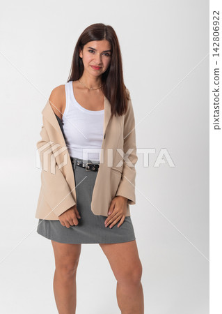 Young woman standing confidently with a subtle smile, wearing a casual yet chic outfit featuring a beige blazer, white tank top, grey mini skirt, and a black belt on a plain background 142806922