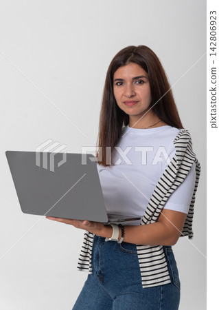 Young adult woman casually dressed, holding a modern laptop and confidently looking forward, representing concepts of technology, connectivity, productivity, and remote work Young adult woman casually dressed, holding a modern laptop and confidently looking forward, representing concepts of technology, connectivity, productivity, and remote work 142806923