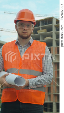 Vertical video. Portrait of an architect or builder in hard hat standing in front of building under construction. Close-up portrait of engineer with blueprint on background of building under Vertical video. Portrait of an architect or builder in hard hat standing in front of building under construction. Close-up portrait of engineer with blueprint on background of building under 142807023