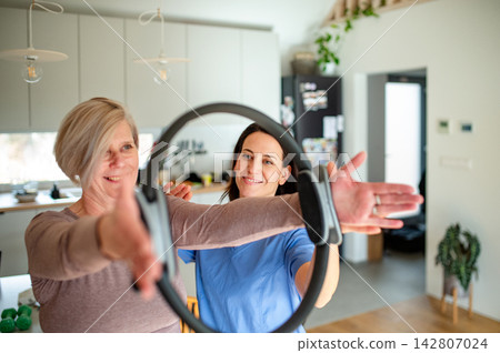 Senior woman performing physiotherapy exercise with ring under physiotherapist supervision at home. Senior woman performing physiotherapy exercise with ring under physiotherapist supervision at home. 142807024