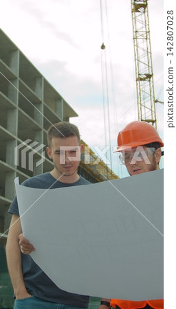 Vertical video. Builder with a male client near building under construction outdoors. Two male builders engineers men, contractor investor worker foreman architect in hard hat discussing drawing Vertical video. Builder with a male client near building under construction outdoors. Two male builders engineers men, contractor investor worker foreman architect in hard hat discussing drawing 142807028