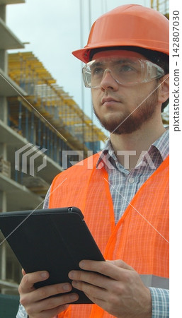 Vertical video. Modern construction engineer or architect in helmet at construction site works with industrial electronic tablet. Close-up portrait of engineer with gadget on background of building Vertical video. Modern construction engineer or architect in helmet at construction site works with industrial electronic tablet. Close-up portrait of engineer with gadget on background of building 142807030