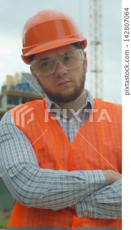 Vertical video. Portrait of an architect or builder in hard hat standing in front of building under construction. Close-up portrait of engineer on background of building under construction. Vertical video. Portrait of an architect or builder in hard hat standing in front of building under construction. Close-up portrait of engineer on background of building under construction. 142807064