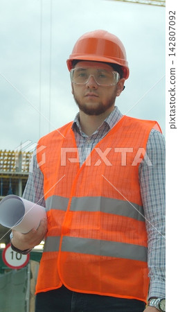 Vertical video. Portrait of an architect or builder in hard hat standing in front of building under construction. Close-up portrait of engineer with blueprint on background of building under Vertical video. Portrait of an architect or builder in hard hat standing in front of building under construction. Close-up portrait of engineer with blueprint on background of building under 142807092