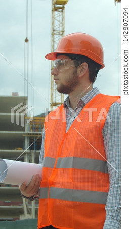 Vertical video. Portrait of an architect or builder in hard hat standing in front of building under construction. Close-up portrait of engineer with blueprint on background of building under Vertical video. Portrait of an architect or builder in hard hat standing in front of building under construction. Close-up portrait of engineer with blueprint on background of building under 142807094