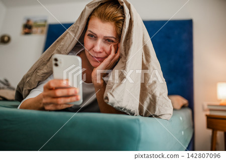 Woman lying under blanket and scrolling on phone. 142807096