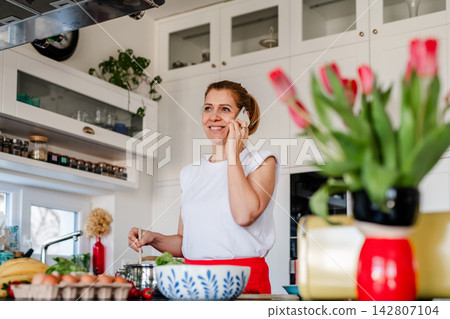 Mature woman making phone call while cooking in kitchen. Mature woman making phone call while cooking in kitchen. 142807104