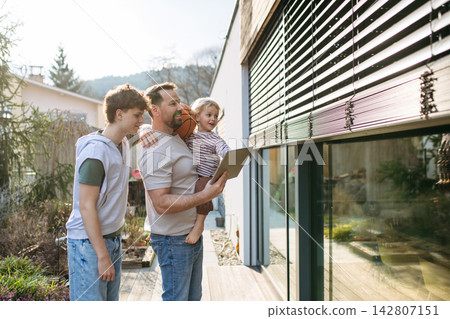 Father controlling smart home exterior blinds using tablet while standing with older and young son. Father controlling smart home exterior blinds using tablet while standing with older and young son. 142807151