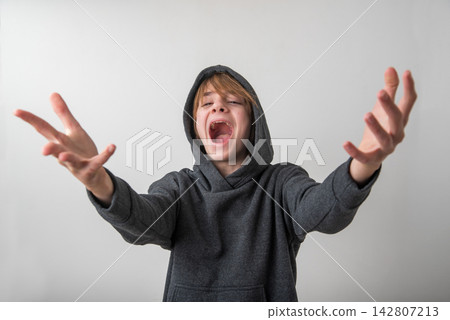 Young boy in a gray hoodie with mouth wide open, arms outstretched and hands reaching forward, expressing intense emotion screaming, anger, frustration, or surprise Young boy in a gray hoodie with mouth wide open, arms outstretched and hands reaching forward, expressing intense emotion screaming, anger, frustration, or surprise 142807213