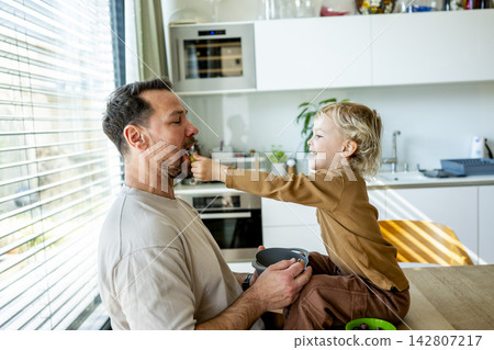 Young child feeding dad healthy snack during playful family moment. Young child feeding dad healthy snack during playful family moment. 142807217