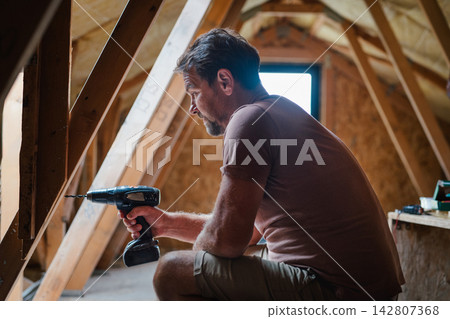 Homeowner doing DIY renovation work with power drill in unfinished attic. Homeowner doing DIY renovation work with power drill in unfinished attic. 142807368