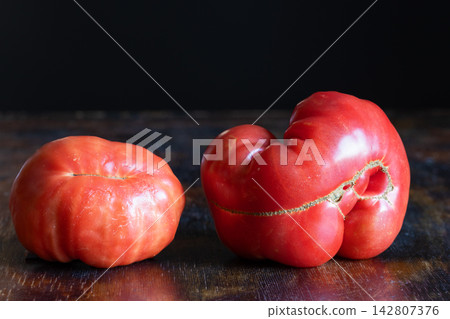 Two ugly, naturally misshapen ripe tomatoes with irregular shapes, deep grooves, folds displayed on rustic wooden table. Contrast between conventional beauty standards and naturally grown vegetables Two ugly, naturally misshapen ripe tomatoes with irregular shapes, deep grooves, folds displayed on rustic wooden table. Contrast between conventional beauty standards and naturally grown vegetables 142807376