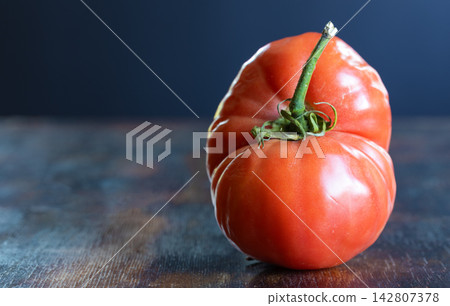 Close-up of naturally misshapen ripe tomato with unusual elongated, lobed shape, deep grooves, uneven contours on rustic wood table. Contrast between conventional produce and naturally imperfect food Close-up of naturally misshapen ripe tomato with unusual elongated, lobed shape, deep grooves, uneven contours on rustic wood table. Contrast between conventional produce and naturally imperfect food 142807378